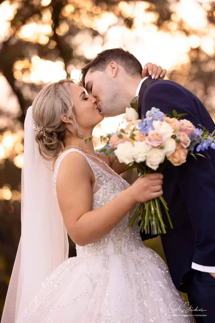 A bride and groom share a kiss at sunset, her bouquet of soft pink and blue flowers in hand