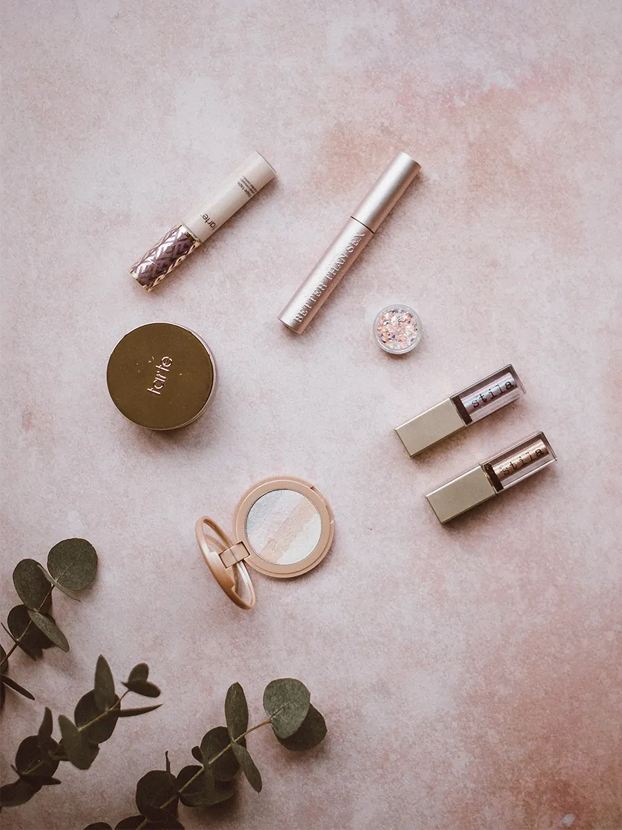 Lipsticks, a compact and gold makeup tools laid out on a blush pink surface with eucalyptus leaves
