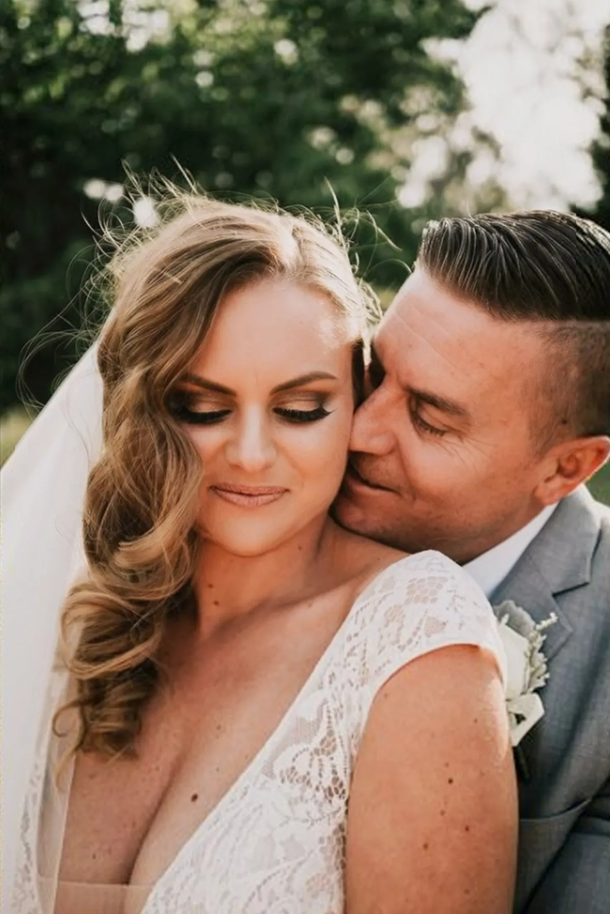 A bride with soft glowing makeup and long brushed-out waves leans into her groom