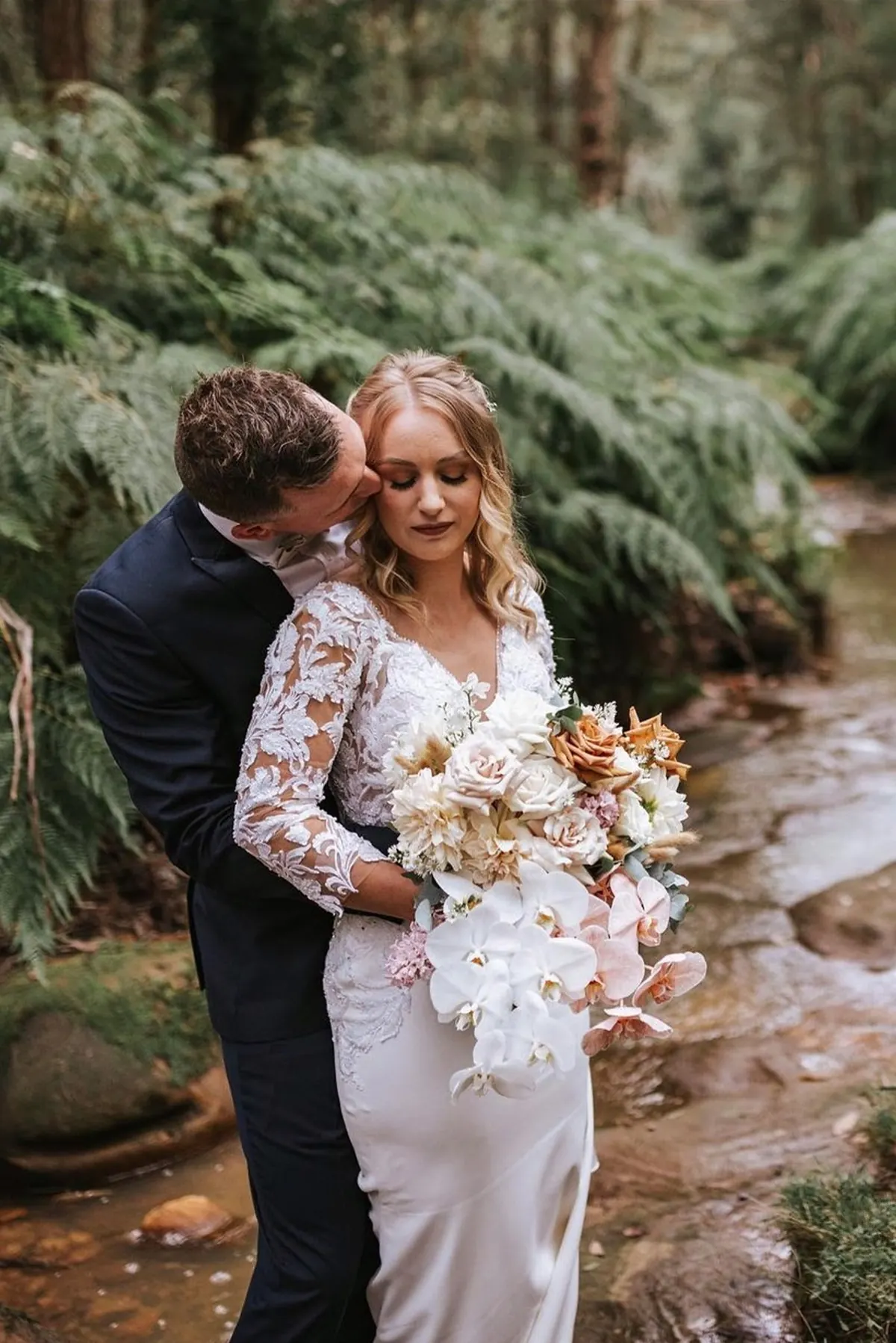 A groom kisses his bride on the cheek beside a fern-lined creek, her hair and makeup by Blown Away